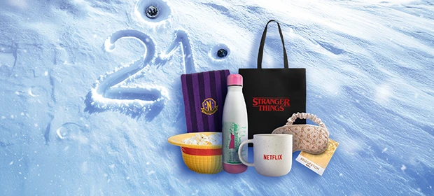  Fanartikel auf Schnee mit Zahl „21“: Stranger-Things-Beutel, Netflix-Tasse, Weiße Flasche mit pinkem Deckel, gelbe Popcornschale, schwarz-lila Schal, Schlafmaske. 