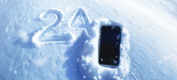  Ein Smartphone, welches zum Teil mit Schneeflocken bedeckt ist, liegt im Schnee. Links daneben die Zahl 24 in den Schnee geschrieben. 
