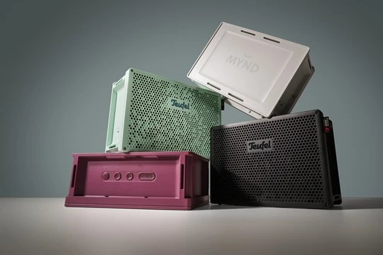  Vier Teufel MYND Lautsprecher in den Farben Warm Black, Warm White, Light Mint und Wild Berry) sind modern arrangiert aufeinandergestapelt und stilvoll ausgeleuchtet vor einer türkisen Wand. 