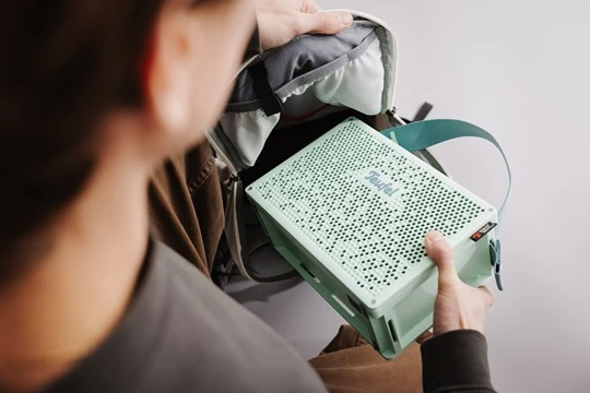  Person packt einen mintgrünen Teufel MYND Lautsprecher mit Trageschlaufe in einen Rucksack. 