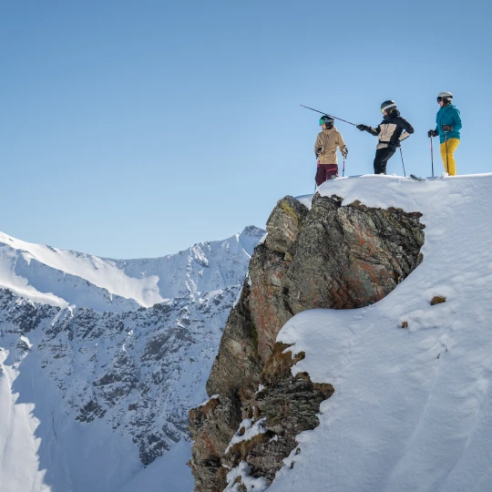  Drei Skifahrer in kompletter Skiausrüstung stehen auf einer schmalen, schneebedeckten Felskante. Einer zeigt mit seinem Skistock auf etwas in der Ferne. Im Hintergrund sieht man hohe, schneebedeckte Berge und blauen Himmel. 