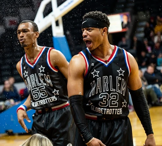  Zwei Harlem-Globetrotters-Spieler in schwarzen Trikots auf dem Spielfeld; der vordere jubelt mit offenem Mund, während feine Partikel durch die Luft fliegen und die Zuschauer unscharf im Hintergrund sind. 