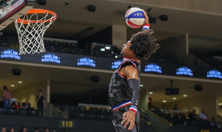  Ein Harlem-Globetrotters-Spieler in schwarzem Trikot springt Richtung Korb und hält den rot-weiß-blauen Ball über dem Kopf zum Dunk; links ist der Ring, im Hintergrund die Arena. 