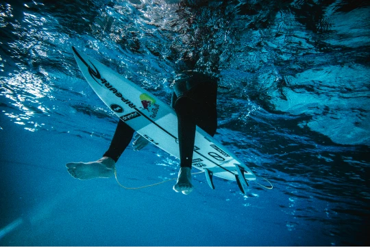 Gewinnspiel Blick von unten auf ein Surfbrett, das sich durch eine Wasserwelle bewegt.