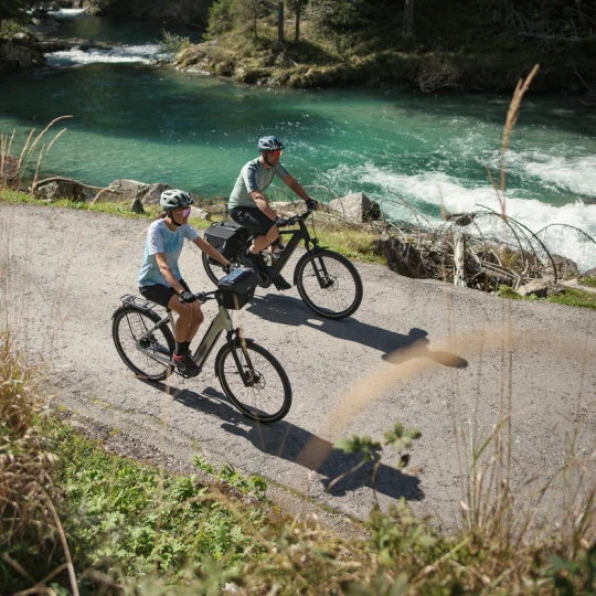 Gewinnspiel FahrradXXL Eine Frau und ein Mann fahren bei Sonnenschein mit E-Bikes an einem türkisblauen Fluss entlang. Beide tragen Fahrradhelme und sportliche Kleidung.