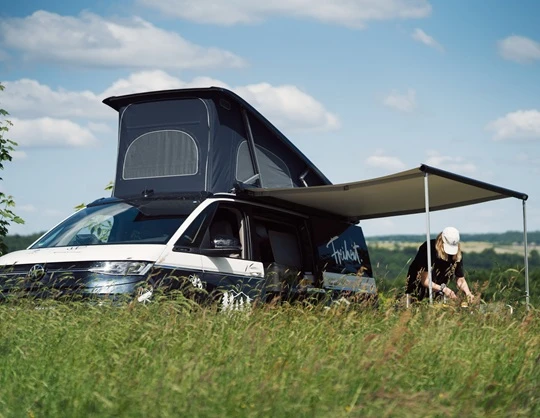  VW Camper mit Markise steht auf einer Wiese, daneben eine Person, die kochen. Es ist ein sonniger Tag und eine weite Landschaft wird im Hintergrund gezeigt. 