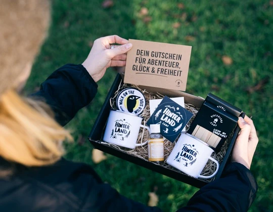  Person hält eine Geschenkbox von Hinterland.camp mit Emaille-Tassen, Aufklebern, Zündhölzern, Patches zum aufbügeln und einer Gutscheinkarte mit der Aufschrift „Dein Gutschein für Abenteuer, Glück & Freiheit“. 