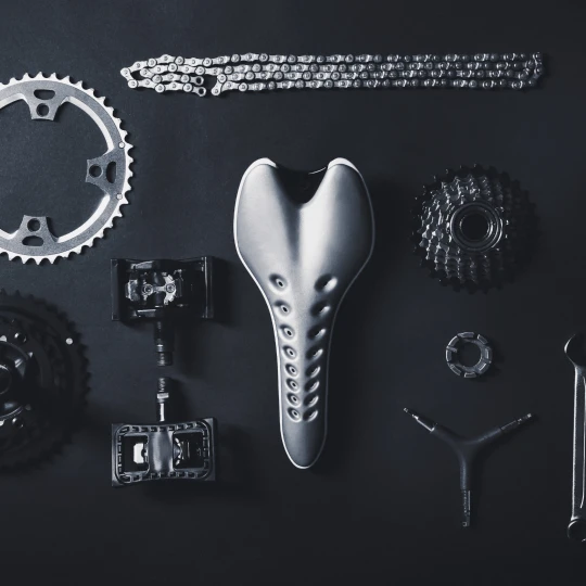 Gewinnspiel FahrradXXL Fahrradteile in Schwarz-Weiß-Optik, darunter ein Sattel, Kette, Pedale, Ritzel, Zahnkranz und Werkzeug, flach angeordnet auf dunklem Hintergrund.