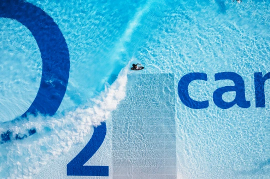 Gewinnspiel Luftaufnahme eines Surfers in einem großen Pool mit O₂-Logo und weißer Wasserbahn.