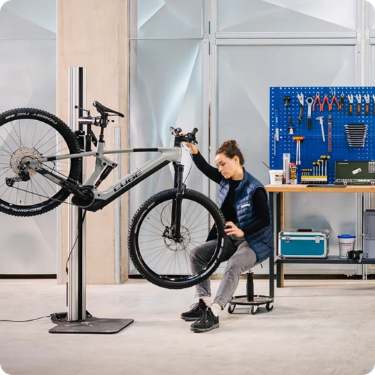  Person arbeitet in einer Werkstatt an einem E-Bike, das in einem Montageständer befestigt ist. Im Hintergrund eine Werkbank mit Werkzeugen und Kiste. 