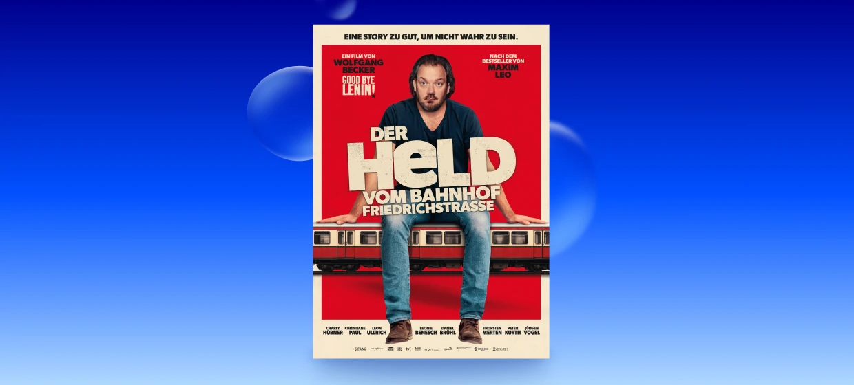  Filmplakat des Films DER HELD VOM BAHNHOF FRIEDRICHST RASSE. Ein Mann sitzt entspannt auf einem Zug, der quer über das Filmplakat verläuft. Hinter ihm ein roter Hintergrund. In der Mitte der große Schriftzug DER HELD VOM BAHNHOF FRIEDRICHSTRASSE. Ganz oben auf dem Plakat: EINE STORY ZU GUT, UM NICHT WAHR ZU SEIN 