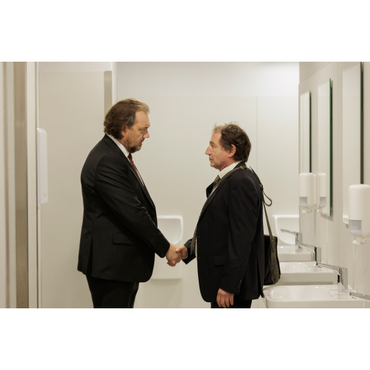 Zwei Männer befinden sich in einem Badezimmer. Sie stehen sich gegenüber und geben sich die Hand . Beide tragen Anzüge und schauen sich ernst an. Im Hintergrund sind Waschbecken und Spiegel zu sehen.