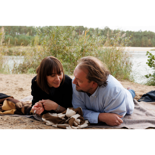 Die Hauptfiguren Micha und Paula liegen gemeinsam auf einer Decke am Seeufer. Sie blicken sich liebevoll an, umgeben von Natur und ruhigem Wasser im Hintergrund.