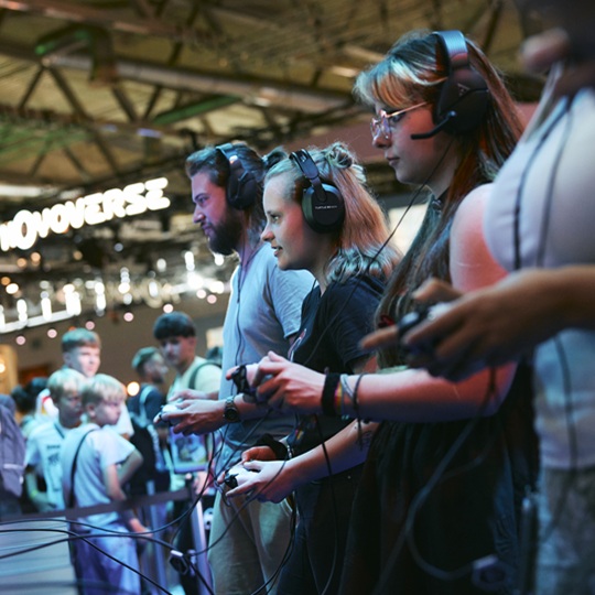 Ein Mann und zwei Frauen mit Gaming-Headsets und Controllern spielen sehr konzentriert und fokussiert ein Videospiel, im Hintergrund erkennt man ein paar Leute die zusehen