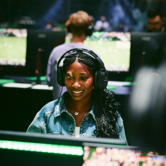 Das Bild zeigt eine junge dunkelhäutige Frau, die einen Gaming-Headset trägt und konzentriert auf einen Computerbildschirm blickt. Sie sitzt in einem Raum mit mehreren anderen Computern im Hintergrund, die ebenfalls Gaming-Aktivitäten zeigen. Die Frau wirkt entspannt, aber auch fokussiert auf das Geschehen auf dem Bildschirm.