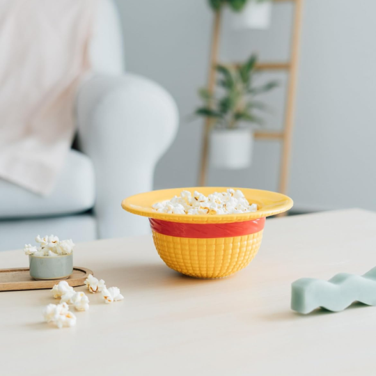 Gelbe Schüssel mit Popcorn auf Tisch, unscharfer Hintergrund mit Sofa und Pflanzen.