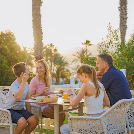 Eine Familie sitzt beim Frühstück im Freien, umgeben von Palmen und Sonnenschein. 