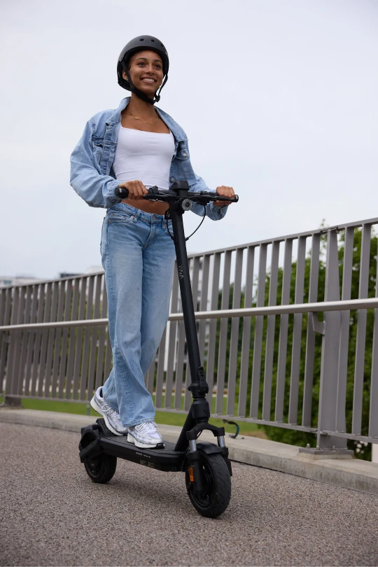  Eine junge Frau mit Helm fährt lächelnd auf dem VMAX VX2 HUB. Im Hintergrund Bäume, eine Grünfläche und Gebäude. 