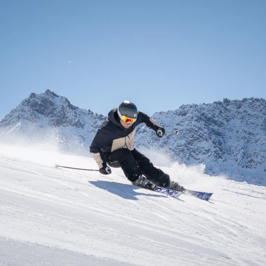  Ein einzelner Skifahrer fährt schnell eine breite Schneepiste hinunter. Er beugt sich sportlich nach vorn und legt sich in eine Kurve. Der Schnee staubt hinter seinen Skiern auf. Der Himmel ist klar und blau und im Hintergrund sieht man hohe, verschneite Berge. 