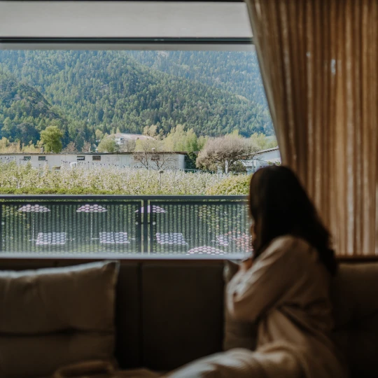  Person sitzt im Innenraum auf einem Sofa und blickt durch ein großes Fenster auf eine Berglandschaft. 