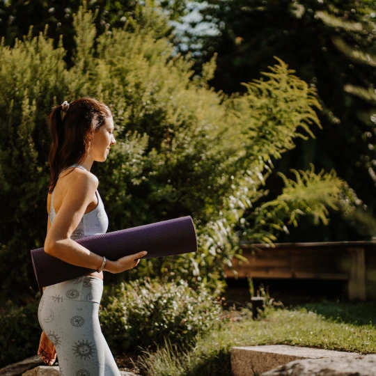  Person geht im Freien mit zusammengerollter Yogamatte durch eine sonnige Gartenanlage mit Bäumen. 