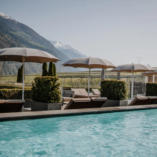  Außenpool mit Liegen und Sonnenschirmen vor Bergkulisse an einem sonnigen Tag. 