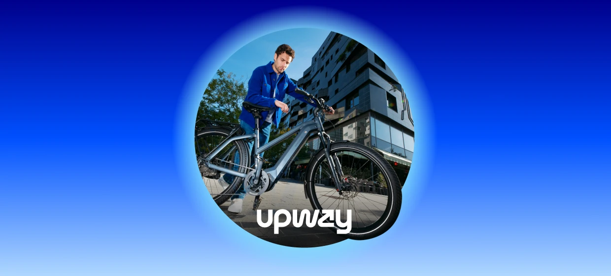  Person steht auf einem gepflasterten Platz und hält ein silbergraues E-Bike. Im Hintergrund moderne mehrstöckige Gebäude mit Glasfassaden und einige Bäume.
