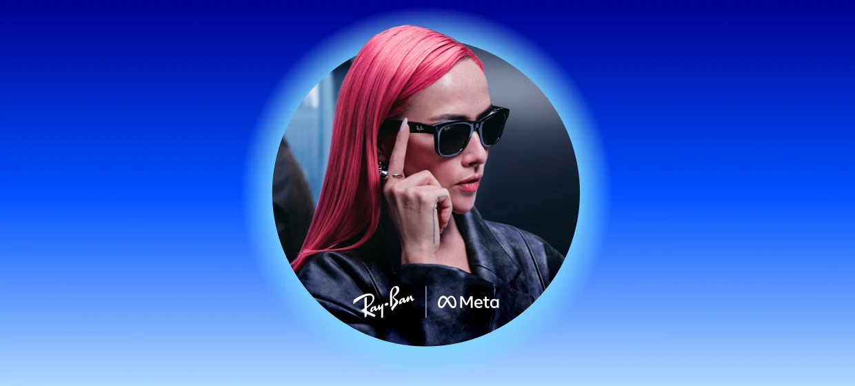  Person mit pinken Haaren trägt schwarze Ray-Ban Meta Wayfarer-Brille und eine dunkle Lederjacke. Mit einem Finger berührt sie den rechten Bügel der Brille. Der Hintergrund ist dunkel und unscharf.
