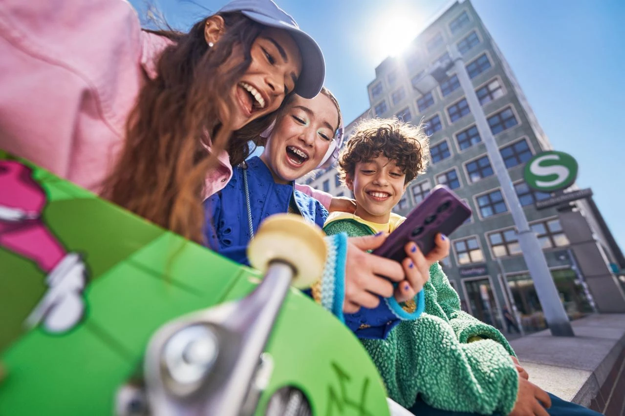  Drei Teenager nutzen im Freien gemeinsam ein Smartphone. Im Vordergrund befindet sich ein grünes Skateboard.
