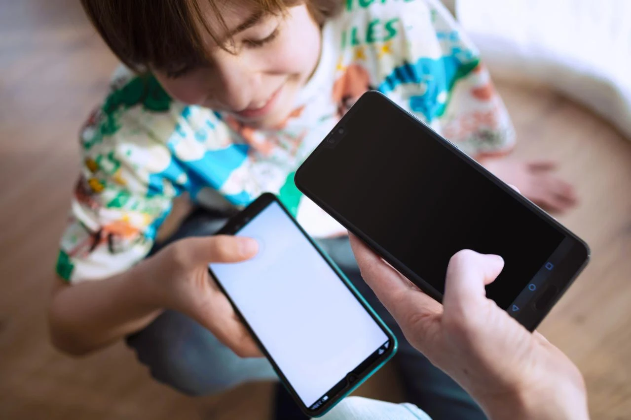  Ein Kind hält ein Smartphone in der Hand und zeigt es einer erwachsenen Person, die ebenfalls ein Smartphone hält.
