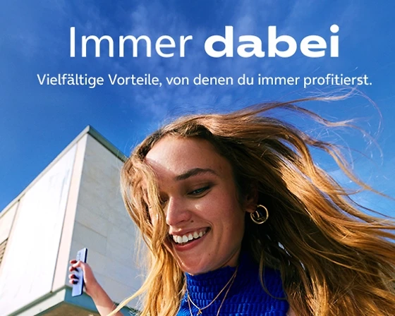 Immer dabei   Eine lachende Frau mit offenen Haaren blickt auf ihr Smartphone. Im Hintergrund ist ein helles Gebäude und blauer Himmel. Überschrift: Immer dabei