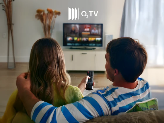  Rückansicht von einer Frau und einem Mann, die auf einen eingeschalteten Fernseher blicken. Der Fernseher steht auf einer weißen Kommode. Über dem Fernseher das O2 TV-Logo. Die Frau hält eine Fernbedienung in der Hand. 