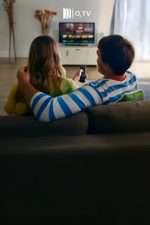  Rückansicht von einer Frau und einem Mann, die auf einen eingeschalteten Fernseher blicken. Der Fernseher steht auf einer weißen Kommode. Über dem Fernseher das O2 TV-Logo. Die Frau hält eine Fernbedienung in der Hand. 