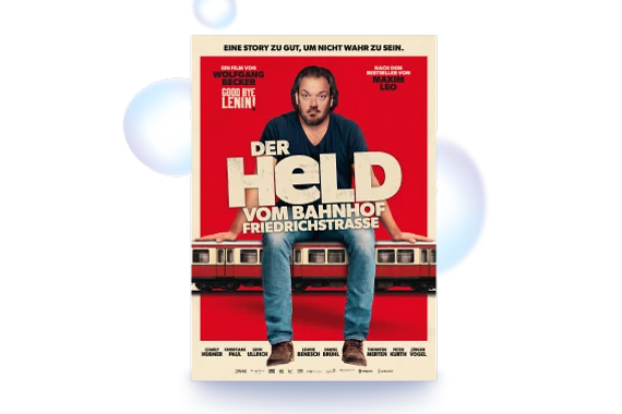  Filmplakat des Films DER HELD VOM BAHNHOF FRIEDRICHST RASSE. Ein Mann sitzt entspannt auf einem Zug, der quer über das Filmplakat verläuft. Hinter ihm ein roter Hintergrund. In der Mitte der große Schriftzug DER HELD VOM BAHNHOF FRIEDRICHSTRASSE. Ganz oben auf dem Plakat: EINE STORY ZU GUT, UM NICHT WAHR ZU SEIN 