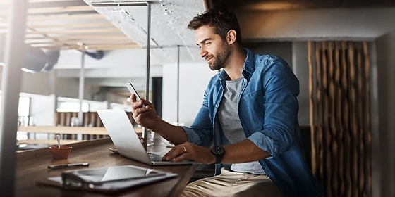 Mann sitzt am Laptop und hält ein Handy in der Hand.